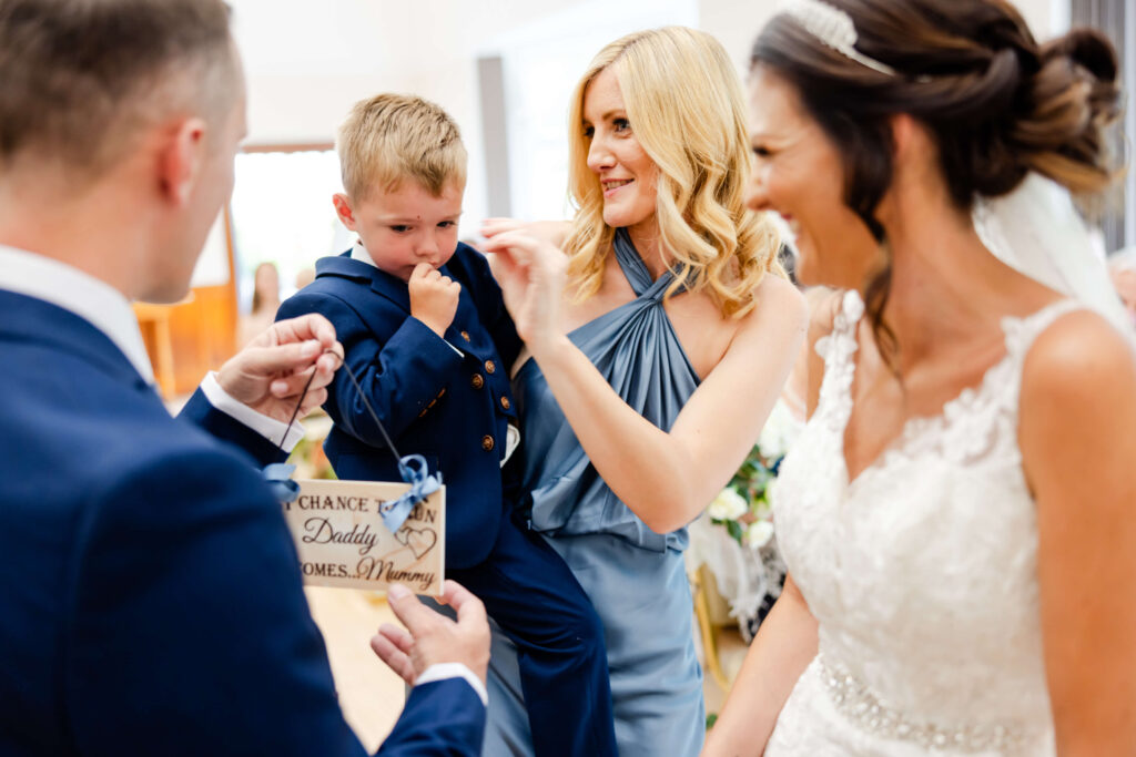 A woman in a blue dress comforts a young boy as a man holds a sign and a bride in a white dress stands nearby at a wedding ceremony. Copyright Somerside Photography Ltd // www.somersidephotography.com