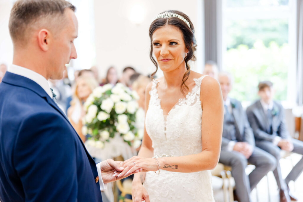 A bride in a white lace dress places a ring on the groom’s finger during a wedding ceremony, with guests seated in the background. Copyright Somerside Photography Ltd // www.somersidephotography.com