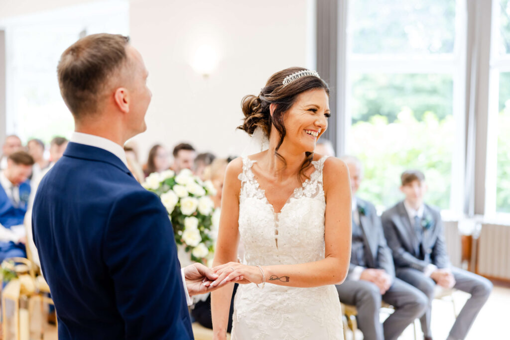 Bride and groom exchange rings and smile during a wedding ceremony, with guests seated in the background. Copyright Somerside Photography Ltd // www.somersidephotography.com