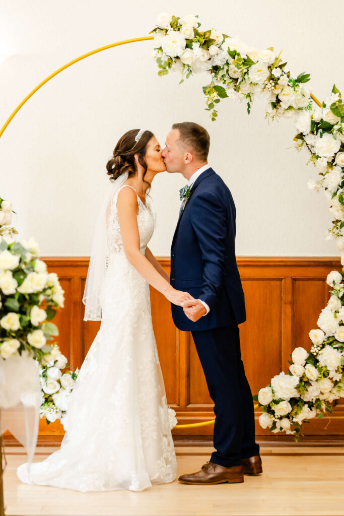 Bride and groom kiss whilst holding hands under a circular floral arch at their wedding ceremony. Copyright Somerside Photography Ltd // www.somersidephotography.com