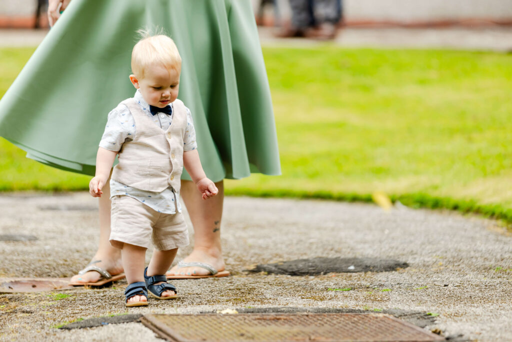 A young toddler in a light-coloured outfit and sandals walks outdoors on a paved path, with an adult in a green skirt standing nearby. Copyright Somerside Photography Ltd // www.somersidephotography.com