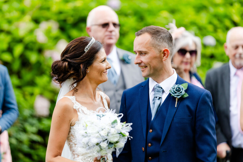 A bride and groom stand close together outdoors, smiling at each other, whilst guests watch in the background. Copyright Somerside Photography Ltd // www.somersidephotography.com