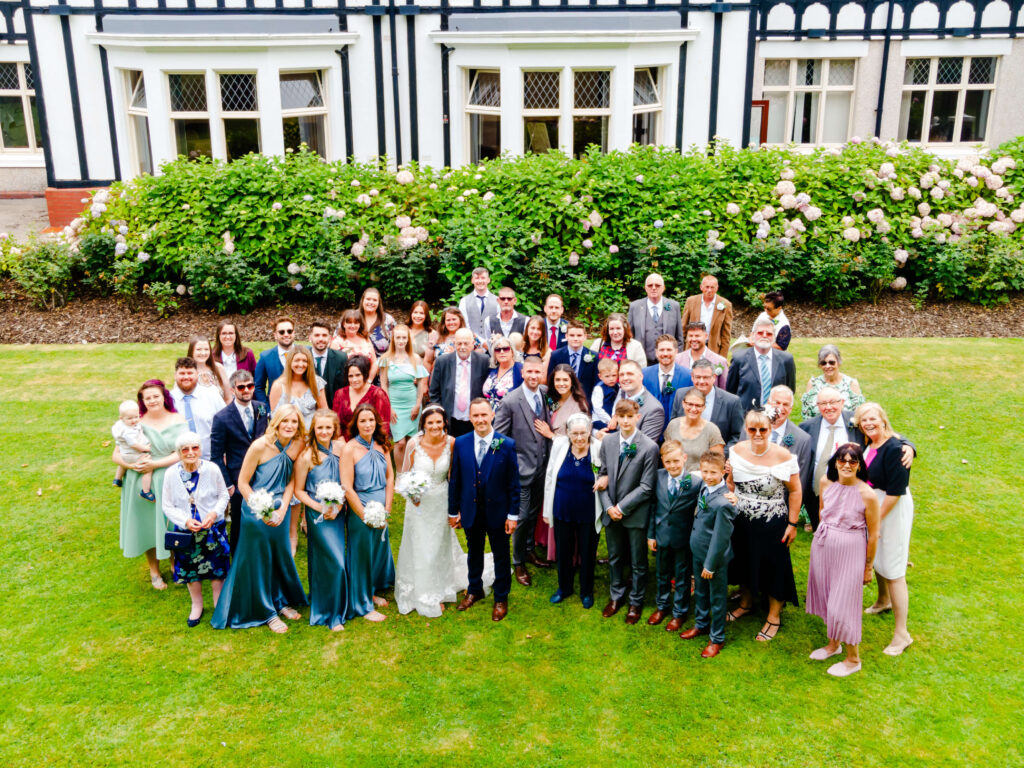 Large group of people, including a bride and groom, pose for a formal wedding photo on a lawn in front of a building with white walls and a hedge of blooming flowers. Copyright Somerside Photography Ltd // www.somersidephotography.com