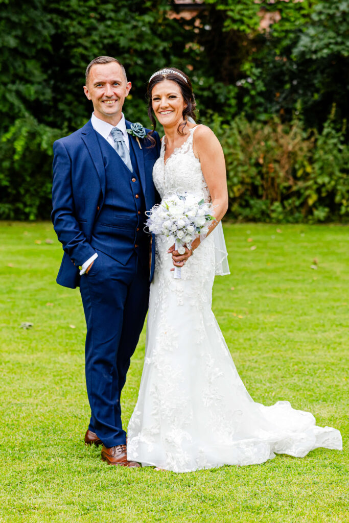A man in a blue suit and a woman in a white wedding dress stand together on grass, smiling at the camera. Copyright Somerside Photography Ltd // www.somersidephotography.com