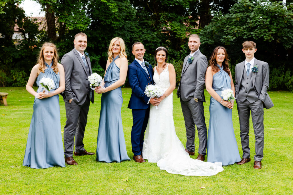 A bride and groom stand together outdoors, flanked by three bridesmaids in blue dresses and three groomsmen in grey suits, all holding bouquets. Copyright Somerside Photography Ltd // www.somersidephotography.com