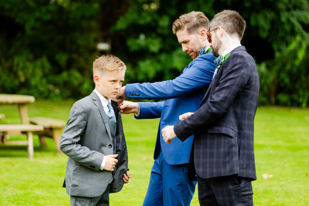 Two men in suits adjust the tie and jacket of a young boy in a suit, standing on grass outdoors with trees in the background. Copyright Somerside Photography Ltd // www.somersidephotography.com