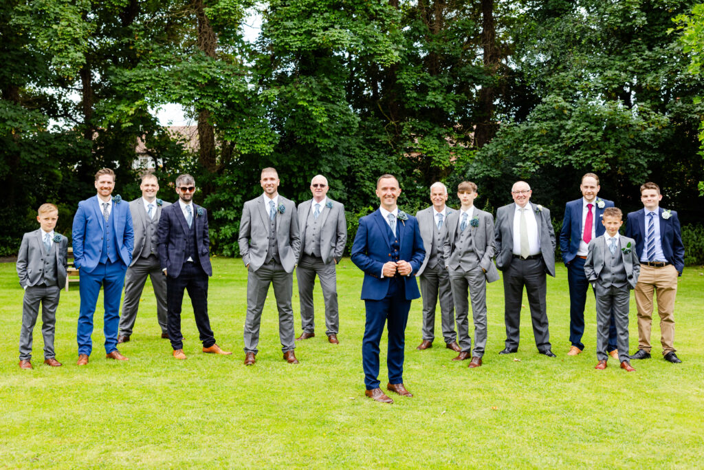 A group of men and boys in suits stand on grass outdoors, with trees in the background. One man in a navy suit stands at the front, holding his hands together and smiling. Copyright Somerside Photography Ltd // www.somersidephotography.com