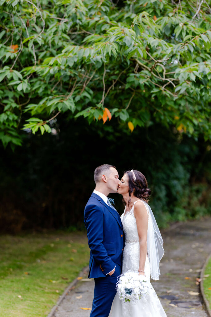 A bride and groom share a kiss outdoors on a pathway beneath green trees, with the bride holding a bouquet and wearing a veil. Copyright Somerside Photography Ltd // www.somersidephotography.com