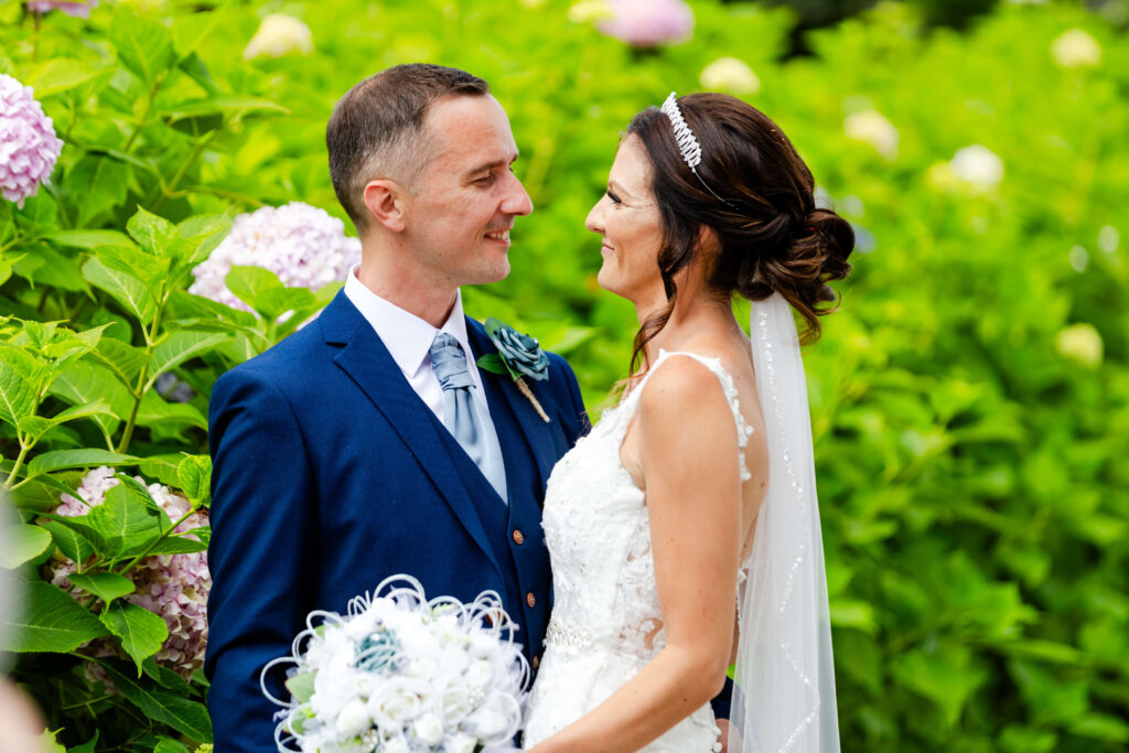 A bride and groom stand facing each other, smiling, surrounded by green foliage and hydrangea flowers. The groom wears a blue suit; the bride wears a white dress and veil. Copyright Somerside Photography Ltd // www.somersidephotography.com