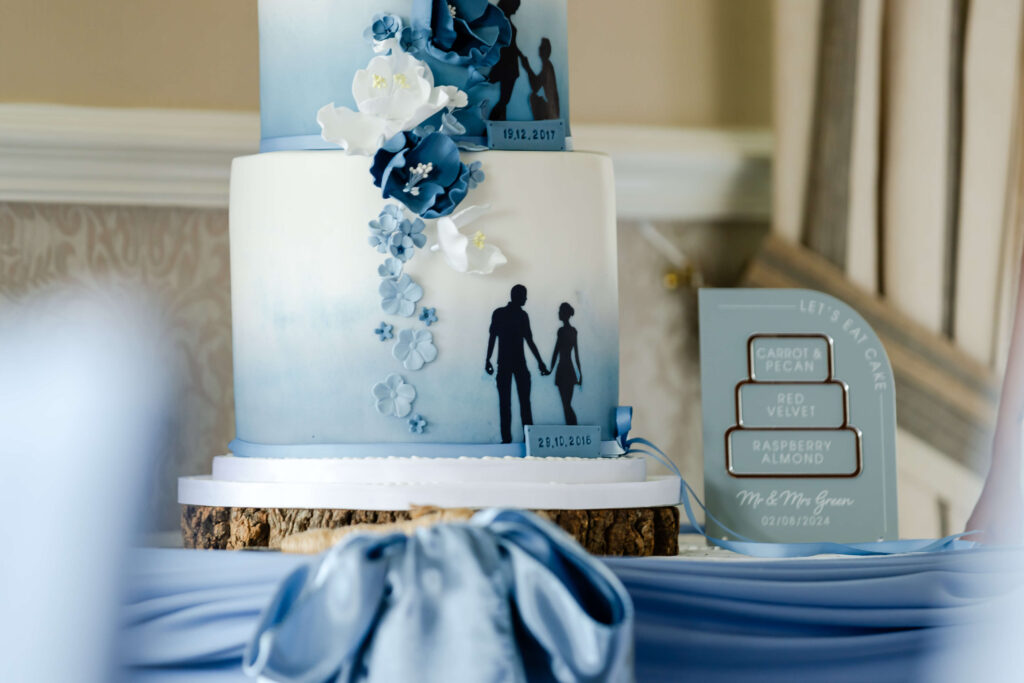 A two-tier blue and white cake with silhouette figures holding hands, decorated with blue and white flowers, sits on a wooden base next to a sign listing cake flavours. Copyright Somerside Photography Ltd // www.somersidephotography.com
