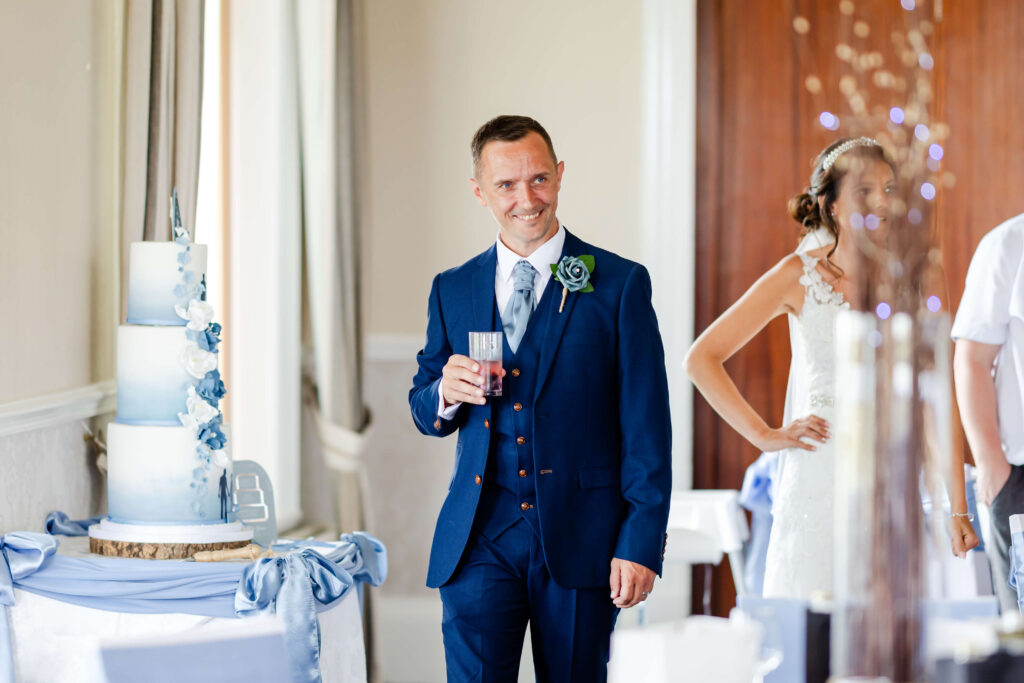 A man in a navy blue suit holding a drink stands indoors near a woman in a wedding dress and a decorated wedding cake. Copyright Somerside Photography Ltd // www.somersidephotography.com