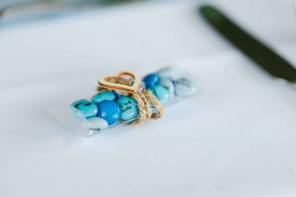 A small clear pouch filled with blue and white sweets, tied with twine and a small wooden heart, placed on a white tablecloth. Copyright Somerside Photography Ltd // www.somersidephotography.com