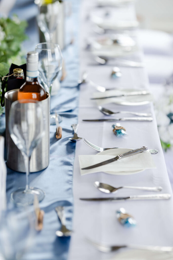 A formal table setting with wine bottles, glasses, cutlery, and serviettes on a white tablecloth with a blue table runner. Copyright Somerside Photography Ltd // www.somersidephotography.com