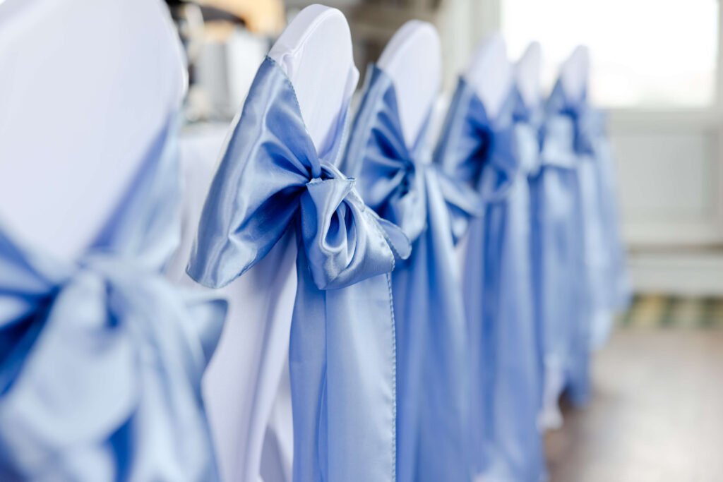 Rows of white chairs are decorated with shiny light blue satin bows tied round the backs, arranged in a line for an event or celebration. Copyright Somerside Photography Ltd // www.somersidephotography.com