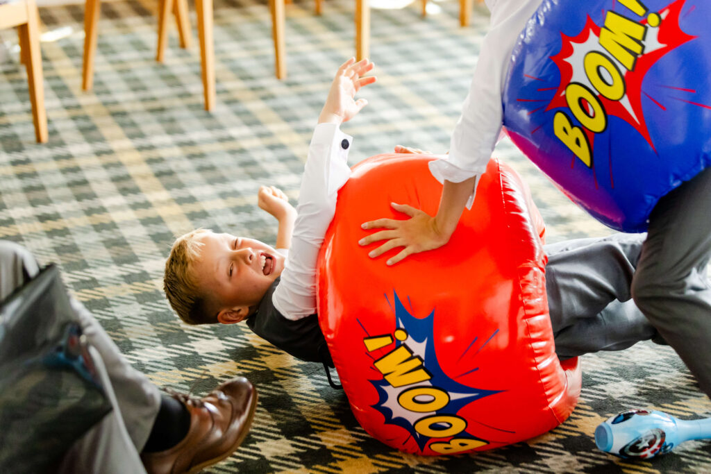 A child in formal clothes lies on a patterned carpet, laughing, whilst another child playfully bumps him with a large inflatable toy that says "BOOM!". Copyright Somerside Photography Ltd // www.somersidephotography.com