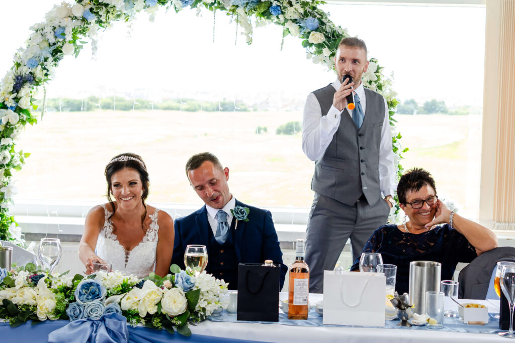 A bride, groom, and two guests sit at a decorated table while a man in a grey waistcoat stands and speaks into a microphone at a wedding reception. Copyright Somerside Photography Ltd // www.somersidephotography.com