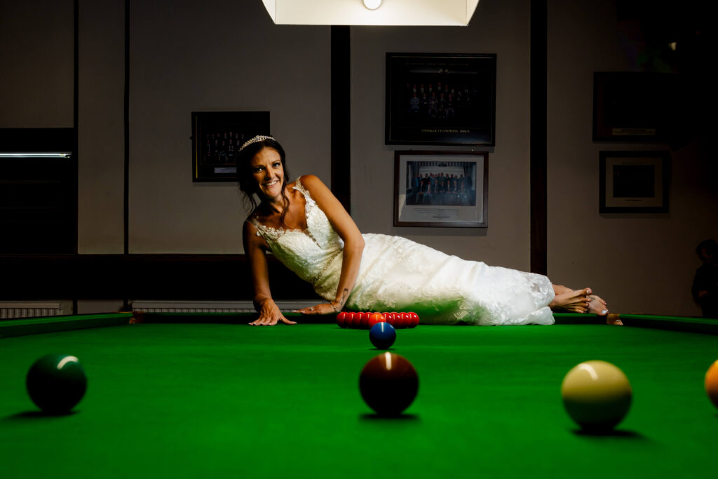 A woman in a wedding dress lies on a snooker table, smiling at the camera, with snooker balls arranged in front of her. Copyright Somerside Photography Ltd // www.somersidephotography.com