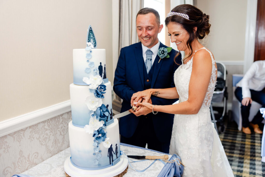 A bride and groom, both smiling, cut a three-tier white and blue wedding cake decorated with flowers and silhouettes of a couple. Copyright Somerside Photography Ltd // www.somersidephotography.com