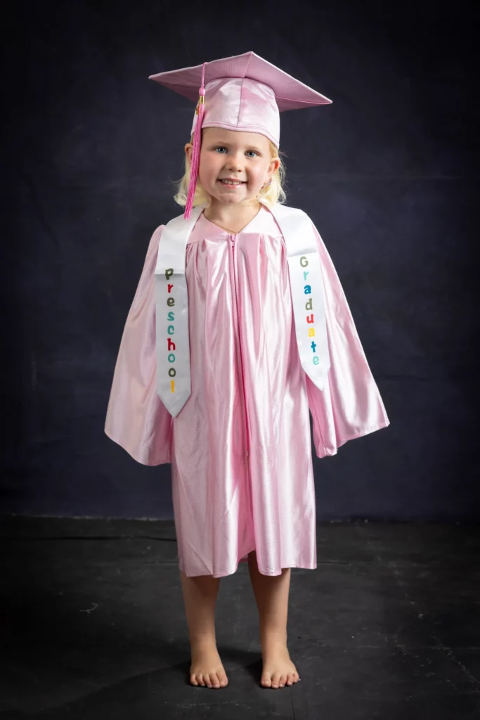Young child in a pink graduation cap and gown, barefoot, stands against a dark background wearing a sash that reads "Preschool Graduate". Copyright Somerside Photography Ltd // www.somersidephotography.com