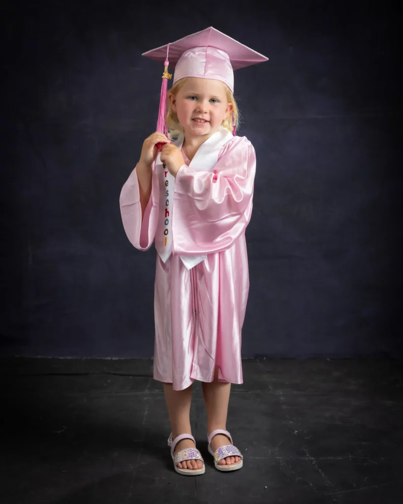 Young girl in a pink graduation cap and gown, holding a tassel, standing in front of a dark background. Copyright Somerside Photography Ltd // www.somersidephotography.com