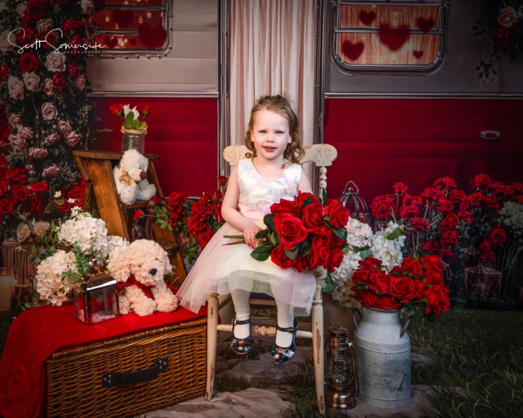 Young girl in a white dress sits on a chair holding red roses, surrounded by flowers, a teddy bear, picnic basket, and vintage decor in front of a red and silver caravan. Copyright Somerside Photography Ltd // www.somersidephotography.com