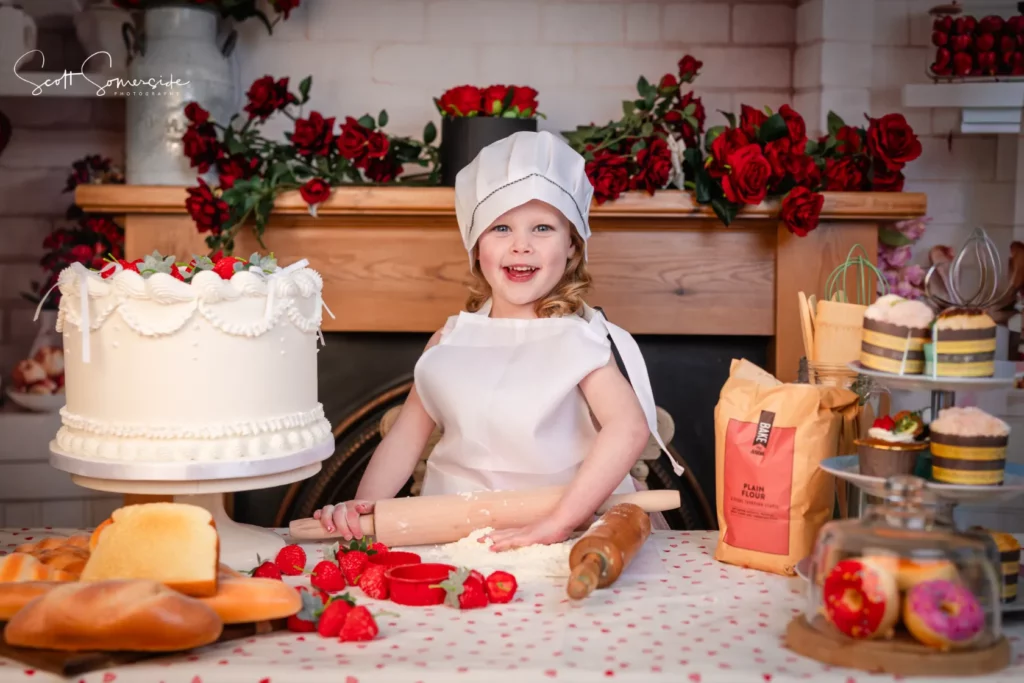 A young child dressed as a baker smiles whilst sitting at a table with baking ingredients, a large decorated cake, and various baked goods in a kitchen decorated with red roses. Copyright Somerside Photography Ltd // www.somersidephotography.com