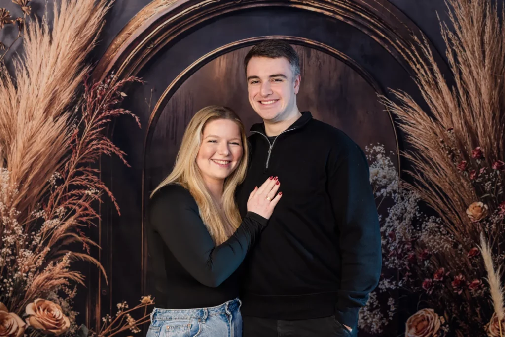 A woman and man stand close together smiling, posing in front of a decorative backdrop with dried plants and flowers. Copyright Somerside Photography Ltd // www.somersidephotography.com