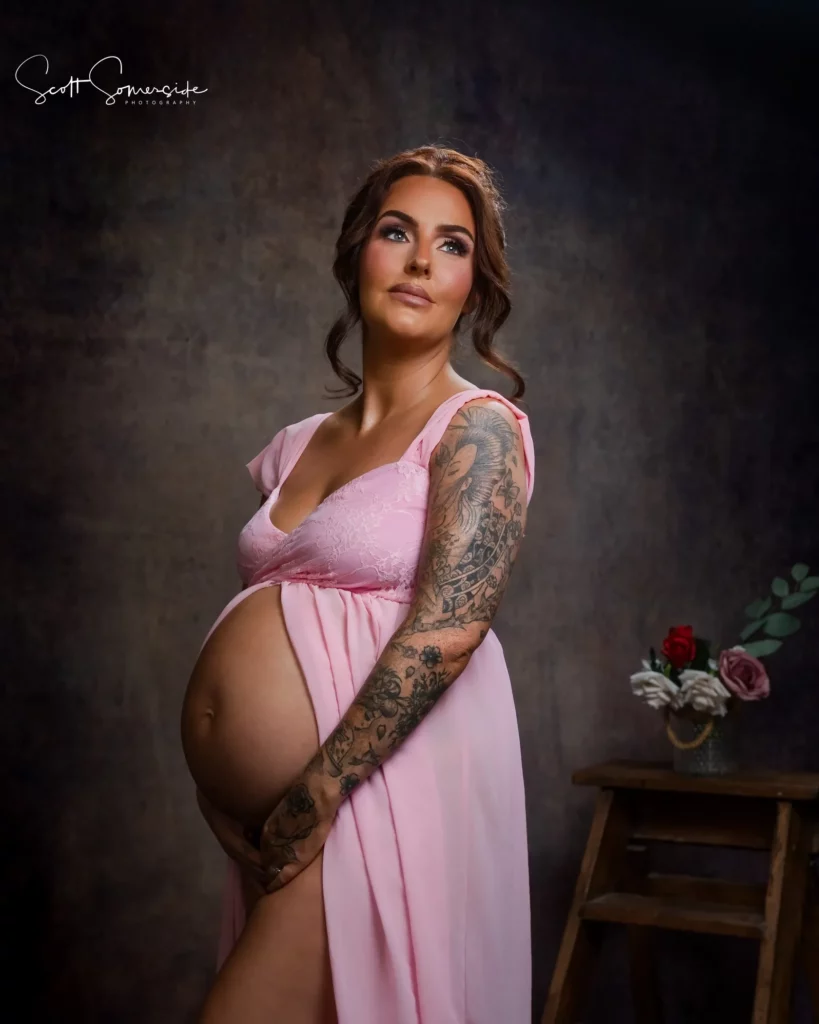 Pregnant woman in a pink dress stands holding her stomach, looking up and to the side, with a stool and flowers in the background. Copyright Somerside Photography Ltd // www.somersidephotography.com