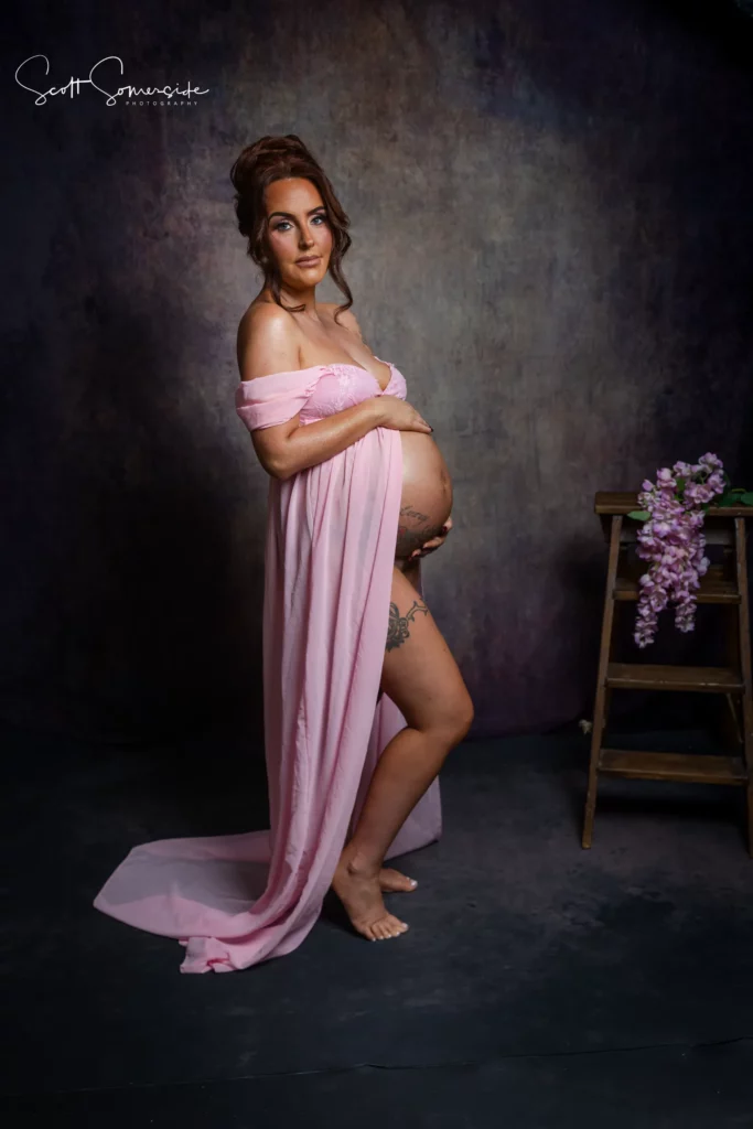 Pregnant woman in a pink dress stands barefoot beside a wooden stool with flowers, holding her bump and looking at the camera against a dark studio backdrop. Copyright Somerside Photography Ltd // www.somersidephotography.com