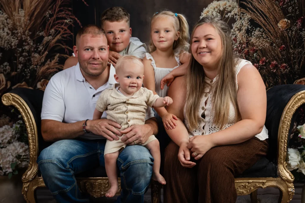 A family of five, including two adults and three young children, sits on a decorative sofa in front of a floral backdrop, posing and smiling for a portrait. Copyright Somerside Photography Ltd // www.somersidephotography.com