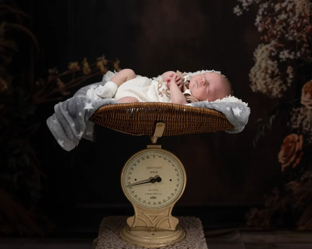 A newborn baby sleeps in a wicker basket lined with a blanket, placed on top of a vintage analogue weighing scale against a dark floral background. Copyright Somerside Photography Ltd // www.somersidephotography.com