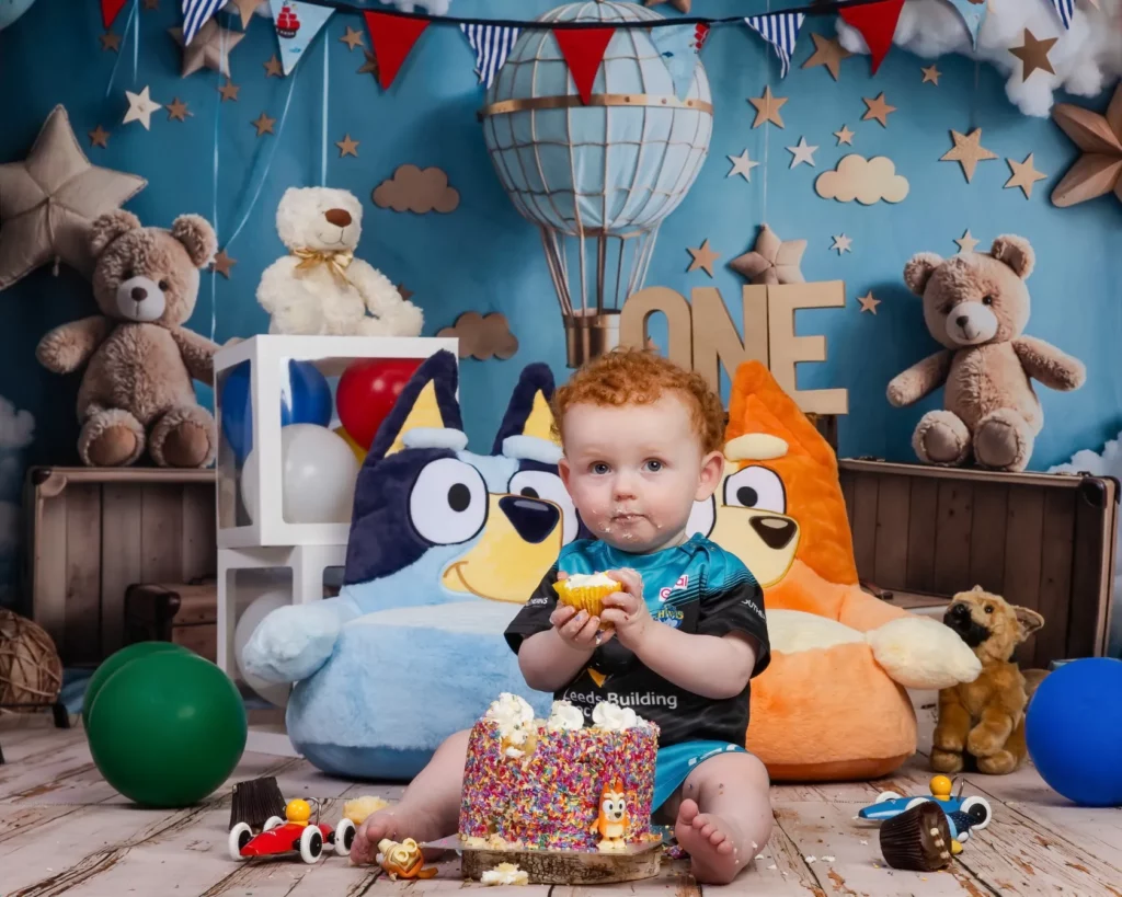 A toddler with red curly hair sits on the floor eating cake at a birthday party, surrounded by soft toys, balloons, and festive decorations with "ONE" in the background. Copyright Somerside Photography Ltd // www.somersidephotography.com