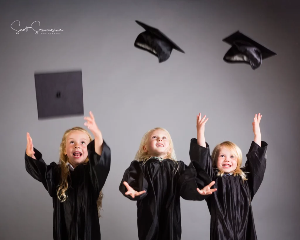 Three young children in graduation gowns throw their caps in the air against a plain background. Copyright Somerside Photography Ltd // www.somersidephotography.com