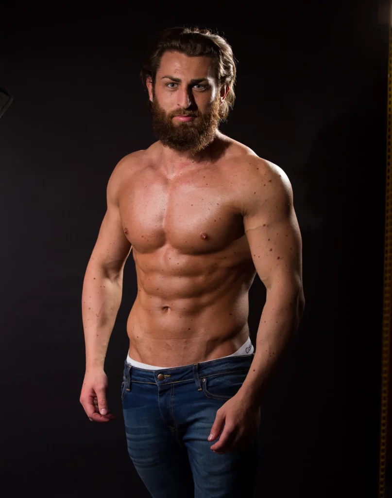 A bearded, bare-chested man with a muscular build stands against a dark background, wearing blue jeans. Copyright Somerside Photography Ltd // www.somersidephotography.com