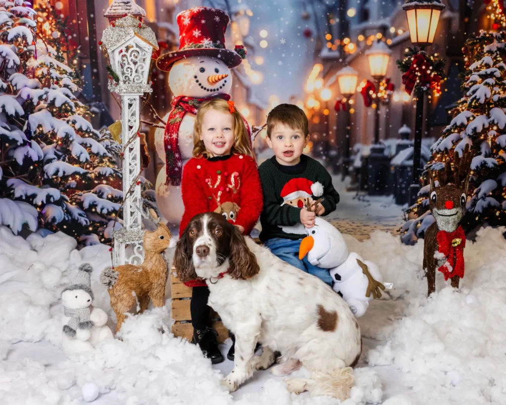 Two children sit on a bench in a snowy, festive scene with Christmas decorations, soft toys, and a brown and white dog in front of them. Copyright Somerside Photography Ltd // www.somersidephotography.com