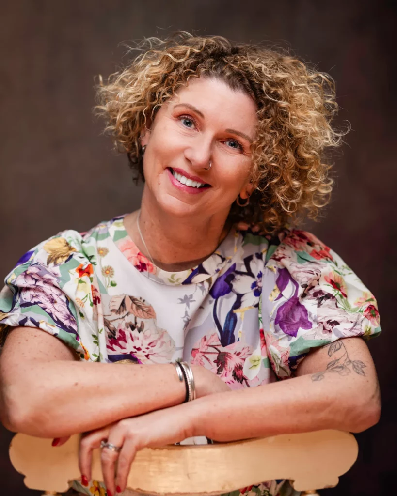 A person with short curly hair smiles while leaning on the back of a chair, wearing a floral patterned top and silver jewellery, with a tattoo visible on one arm. Copyright Somerside Photography Ltd // www.somersidephotography.com