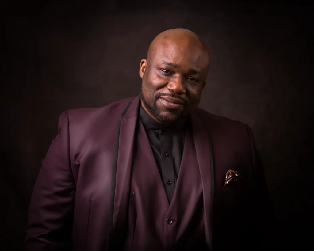 A man in a dark maroon suit and black shirt stands against a dark background, looking at the camera with a slight smile. Copyright Somerside Photography Ltd // www.somersidephotography.com